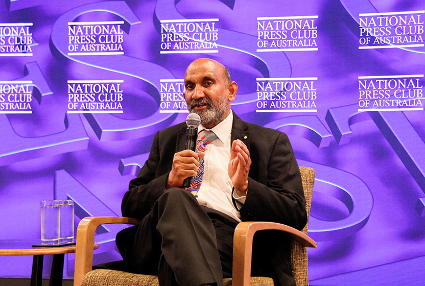 Professor Jagadish is wearing a suit and sitting in a chair while speaking into a microphone. Behind him is a purple background with the words 'National Press Club of Australia'.